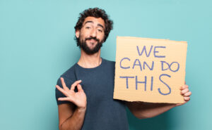 Uśmiechnięty mężczyzna trzymający karton z napisem „We can do this” na niebieskim tle, pokazujący gest OK – symbol pozytywnego nastawienia i zaangażowania
