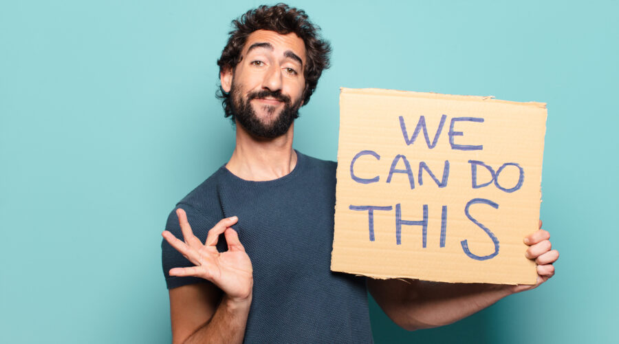 Uśmiechnięty mężczyzna trzymający karton z napisem „We can do this” na niebieskim tle, pokazujący gest OK – symbol pozytywnego nastawienia i zaangażowania